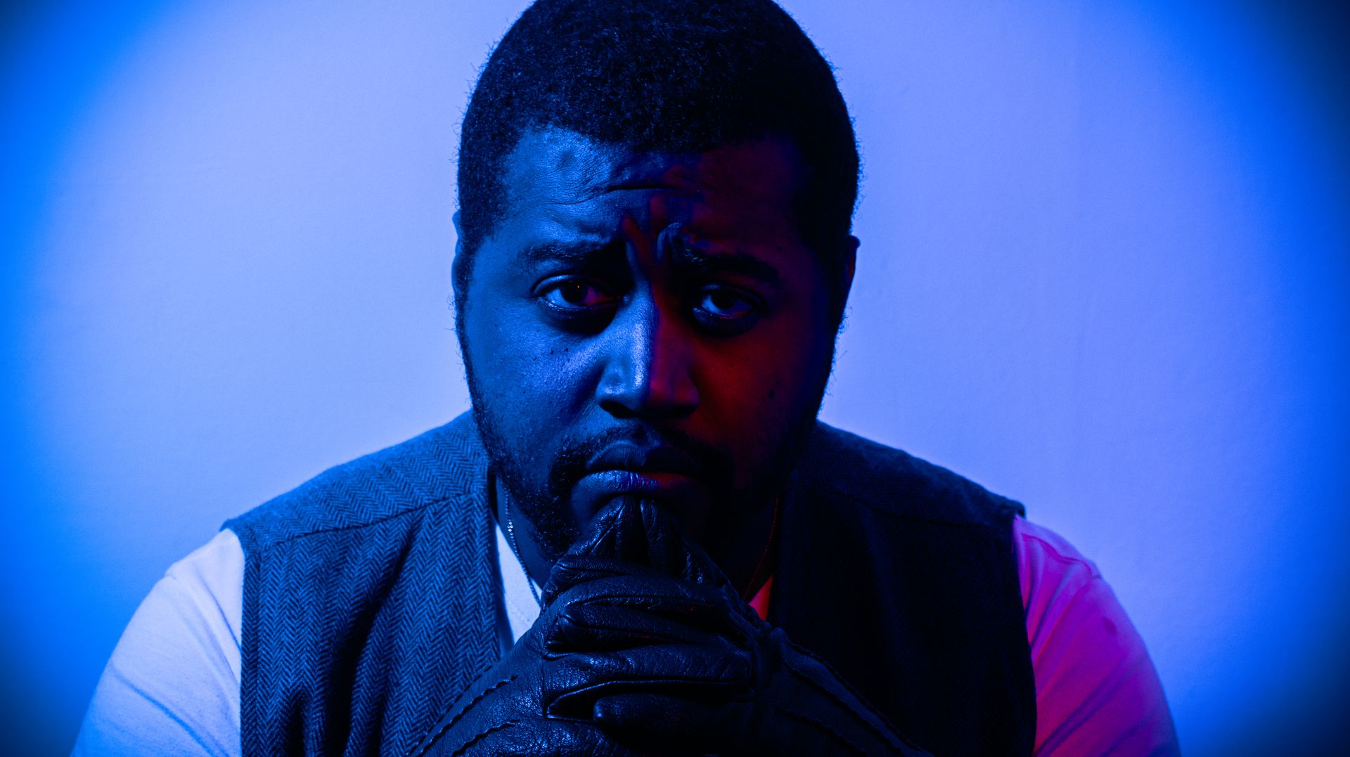 a man in a suit looking troubled with leather gloves, and red and blue lighting shining on him